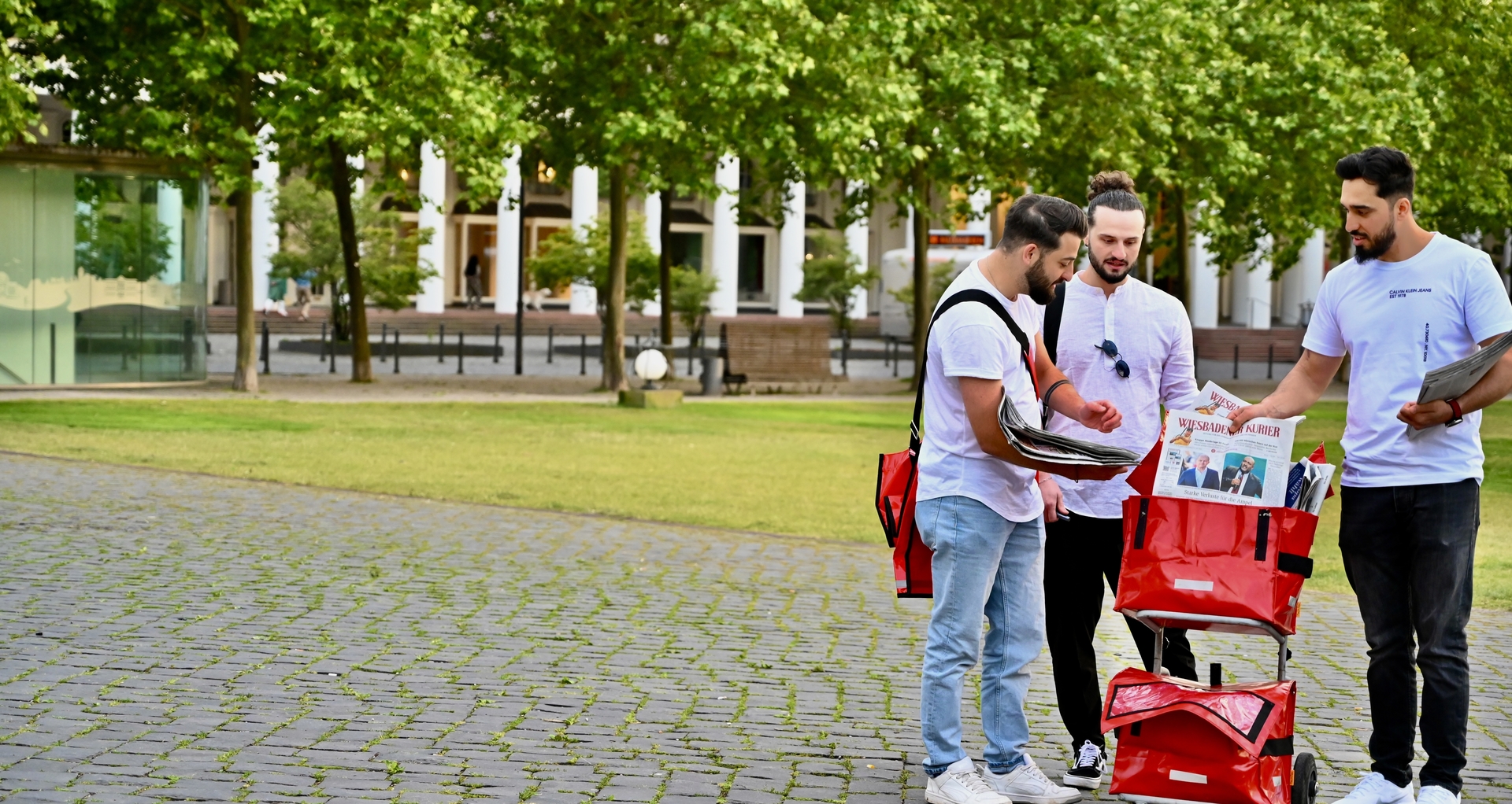 Drei Zusteller mit Zeitungswagen und Zeitungen auf einem großen Platz mit Bäumen im Hintergrund