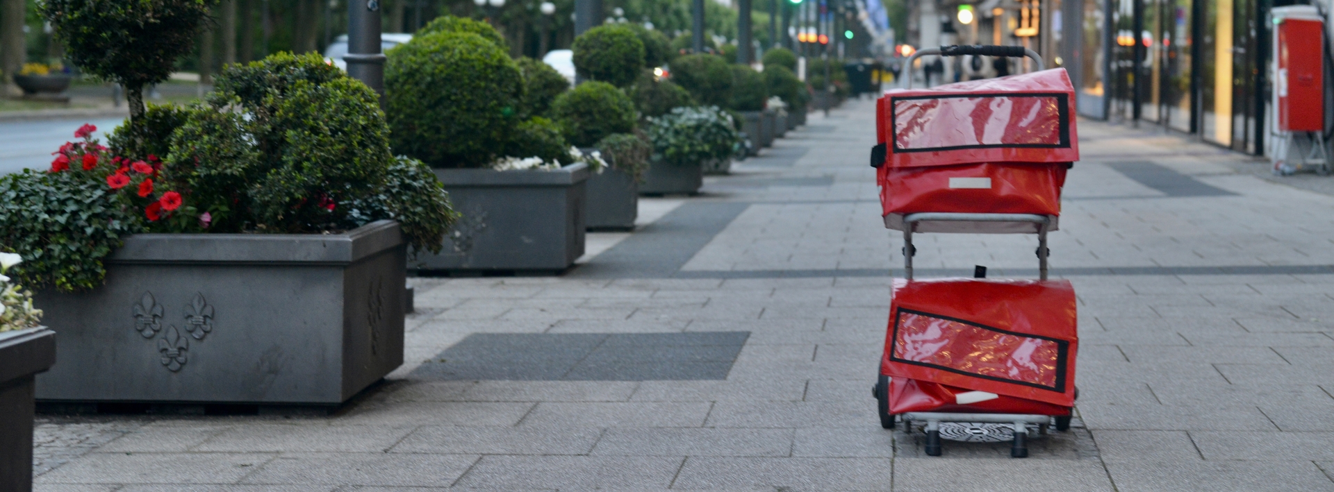 Roter Zeitungswagen auf einem Gehweg mit Geschäften rechter Hand und Blumenkübeln linkerhand